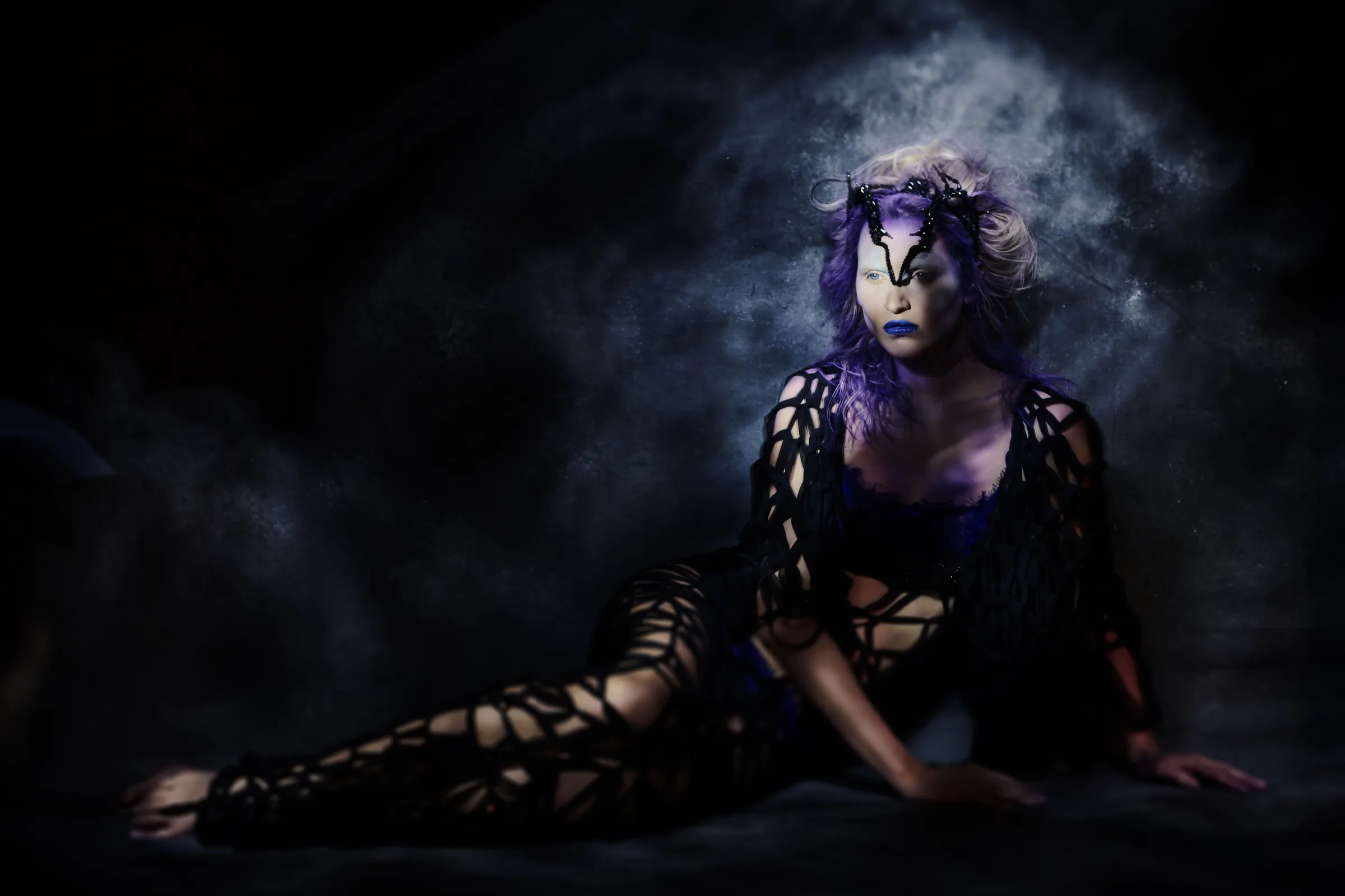 A seated figure in black emerges from violet light and smoke.